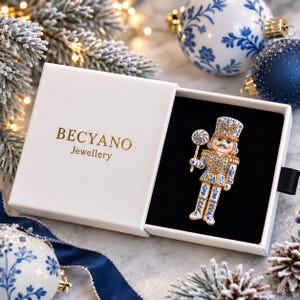 May include: A nutcracker brooch with blue and white floral patterns and gold accents, held in a white rectangular box with the gold text BECYANO Jewellery printed on the lid. The brooch features a soldier figure holding a lollipop-shaped staff.