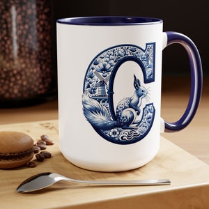 Op de afbeelding: Witte keramische mok met een blauw interieur en handvat. De mok heeft een blauw-wit bloemmotief met een eekhoorn en de letter "C". De mok staat op een houten oppervlak.