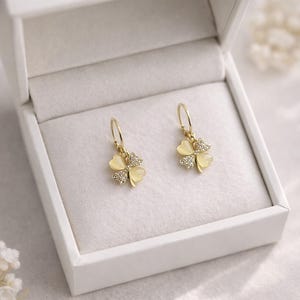 Peut inclure: Une paire de boucles d'oreilles dorées, chacune ornée d'un charm en forme de trèfle à quatre feuilles. Les feuilles de trèfle sont jaune pâle, avec des cristaux clairs scintillants au sommet de chaque feuille. Les boucles sont dans un écrin blanc.