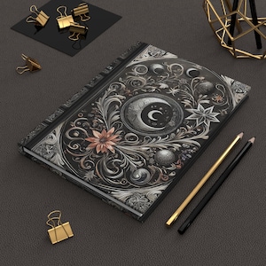 Op de afbeelding: Een zwart notitieboek met een zilveren en gouden bloemenontwerp met een halve maan en sterren. Het notitieboek heeft een textuur.