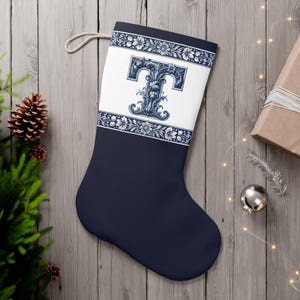 Op de afbeelding: Marineblauwe kerstsok met een wit paneel met een decoratieve blauwe letter "T". De boven- en onderkant van het witte paneel hebben een bloemenrand. Bovenop is een beige lus bevestigd.