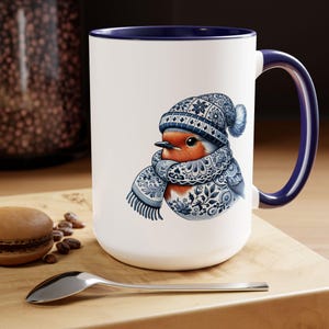 May include: White ceramic mug with a blue rim and handle. The mug features a colorful illustration of a robin wearing a blue and white patterned hat and scarf.