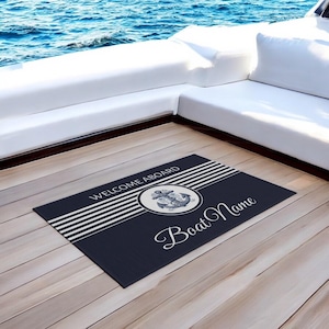 Pode incluir: Um tapete de boas-vindas azul marinho com listras brancas e um círculo branco contendo um desenho de âncora azul. O texto "Welcome Aboard" e "Boat Name" está impresso no tapete.