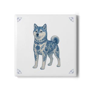 May include: Square ceramic tile featuring a Shiba Inu dog illustration. The dog is depicted in blue and white, with intricate floral and leaf patterns. Decorative blue flourishes are in each corner of the white tile.