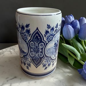 May include: A white ceramic vase with blue floral and landscape designs. The vase has a cylindrical shape with a slightly flared rim. The blue designs include flowers, foliage, and scenes. A bouquet of blue tulips is in the background.