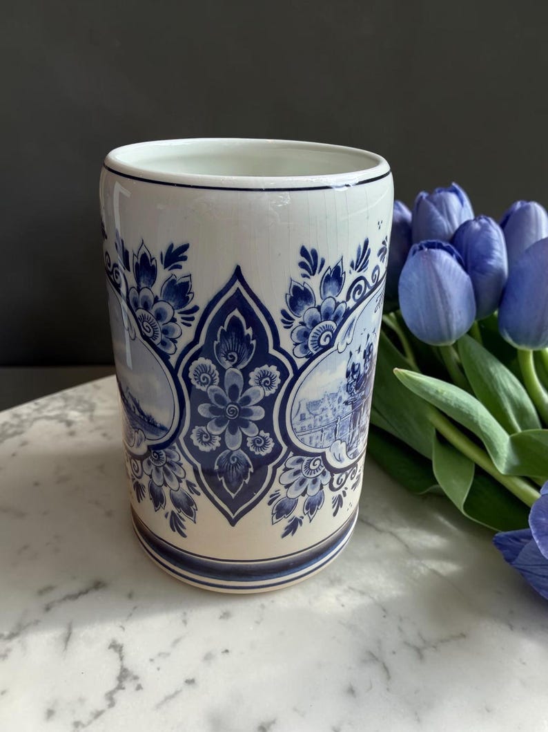 May include: A white ceramic vase with blue floral and landscape designs. The vase has a cylindrical shape with a slightly flared rim. The blue designs include flowers, foliage, and scenes. A bouquet of blue tulips is in the background.