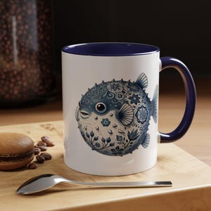 May include: White ceramic mug with a blue interior and handle. The mug features a detailed illustration of a blue pufferfish with floral and swirl patterns. A spoon and a pastry are in the foreground, resting on a wooden surface.