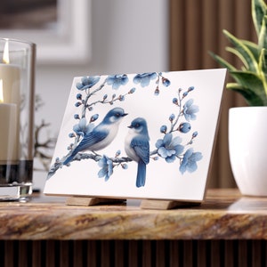 Könnte beinhalten: Eine weiße Keramikfliese mit einem blauen und weißen Blumendesign, auf dem zwei blaue Vögel auf einem Ast sitzen. Die Fliese steht auf einem Holzständer.