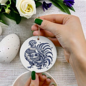 Op de afbeelding: Een witte keramische ornament met een blauw en wit haanontwerp. De haan is versierd met ingewikkelde patronen en heeft een gouden touwtje om op te hangen.