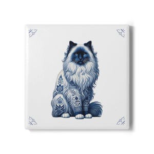 Puede incluir: Un azulejo de cerámica azul y blanco con una ilustración detallada de un gato de pelo largo con ojos azules. El gato está sentado con las patas cruzadas y tiene un patrón decorativo en su pelaje.