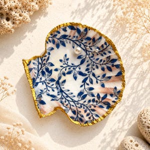 Op de afbeelding: Een decoratieve sint-jakobsschelp met een witte basis, een blauw botanisch bladpatroon en een metallic goudkleurige rand. De binnenkant van de schelp is versierd met drie kleine witte bolvormige kralen in een groepje.