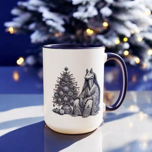 Op de afbeelding: Een witte keramische mok met een blauwe rand heeft een zwart-witte illustratie van een personage uit Star Wars dat naast een gedecoreerde kerstboom staat. De mok heeft een blauwe handgreep.