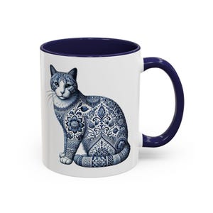 Może przedstawiać: Biały kubek ceramiczny z niebieskim wnętrzem i uchwytem. Kubek przedstawia niebiesko-białą ilustrację kota z kwiatowym wzorem.