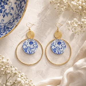 Op de afbeelding: Een paar goudkleurige oorbellen met een gestructureerde gouden schijf verbonden aan een grotere gouden ring. Binnenin elke ring hangt een ronde witte bedel versierd met een blauw bloemmotief. De oorbellen hebben standaard haaksluitingen.