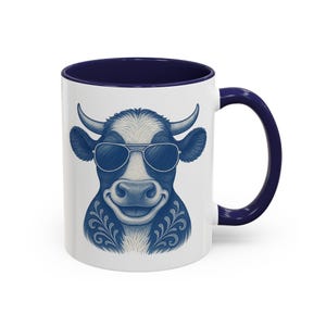 Op de afbeelding: Witte keramische mok met een marineblauwe binnenkant en handvat. De mok heeft een lachende koe met een zonnebril, met een blauw-wit ontwerp. De koe heeft hoorns en decoratieve bloemmotieven. Een leuke en unieke koffiemok.