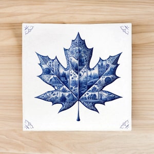 Op de afbeelding: Een blauw en wit keramisch esdoornbladontwerp met ingewikkelde details. Het blad is gemaakt van kleinere tegels die een patroon van huizen, bomen en andere ontwerpen creëren.