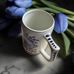 May include: A white ceramic mug with blue floral patterns and a distinctive handle. The mug is set against a backdrop of blue tulips with green stems. The mug's design features a detailed illustration in blue.