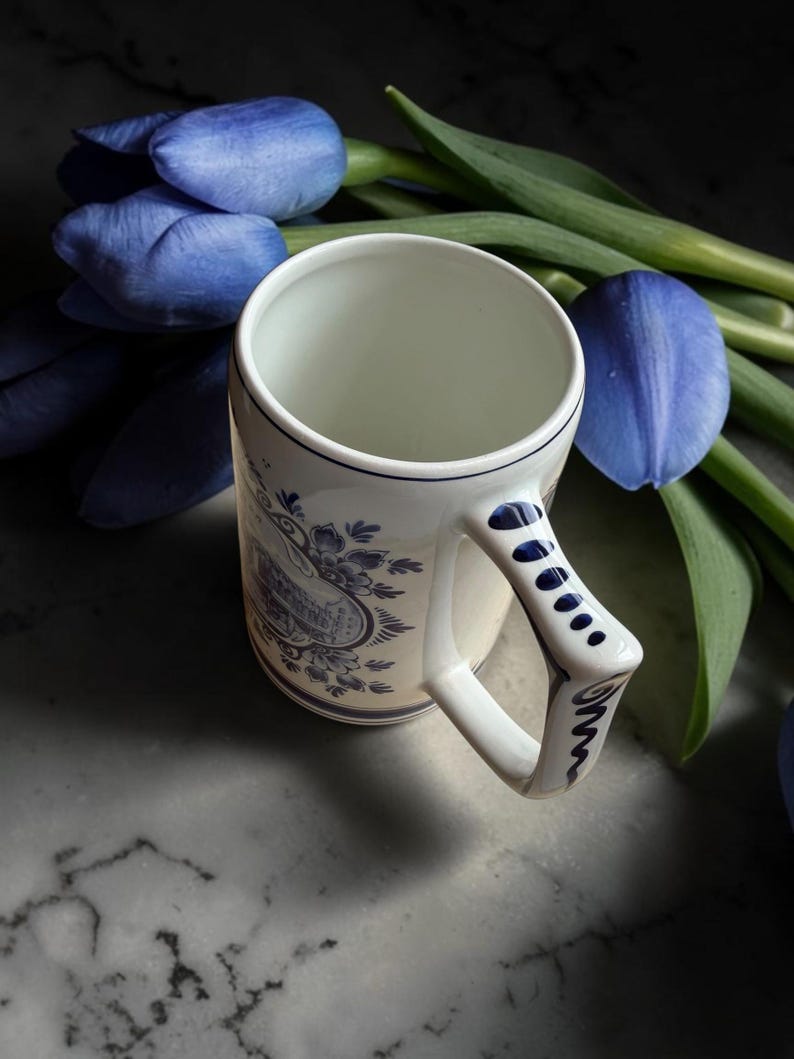 May include: A white ceramic mug with blue floral patterns and a distinctive handle. The mug is set against a backdrop of blue tulips with green stems. The mug's design features a detailed illustration in blue.