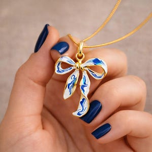 May include: A gold-colored necklace with a bow pendant. The bow is white with blue and gold accents. The necklace is held by a person with dark blue nail polish.