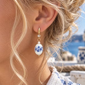 Könnte beinhalten: Ein goldfarbener Ohrhänger mit einem klaren, runden Kristall, einer kleinen weißen Perle und einem ovalen, weißen Keramik-Anhänger, der mit einem blauen Blumenmuster verziert ist. Der Ohrring hängt an einem goldfarbenen Haken und wirkt filigran.