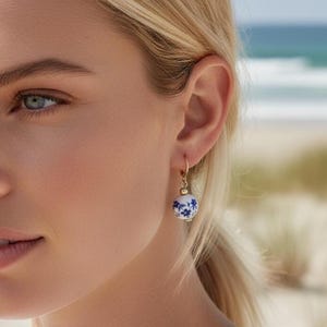 Könnte beinhalten: Goldfarbene Creolen mit einer runden, weiß-blauen Perle mit floralem Muster. Die Perle hat ein zartes blaues Blumenmuster. Die Ohrringe sind an einem Ohr zu sehen, mit einer Strandszene im Hintergrund.