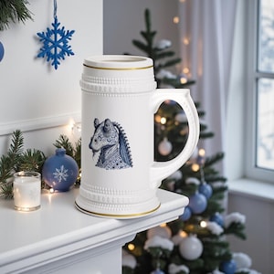 May include: A white ceramic beer stein with a gold rim and handle. The stein features a blue and white illustration of a creature. The stein is decorated with embossed details. The stein is displayed on a white surface with Christmas decorations in the background.