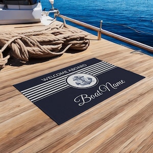 以下が含まれることがあります： 白のストライプと、青い錨と「Welcome Aboard」の文字が入った白い円が描かれたネイビーブルーのウェルカムマット。円の真下に「Boat Name」の文字が白い筆記体で書かれています。