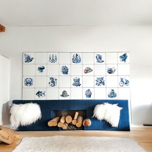 May include: A blue and white tiled wall with a variety of images, including a bird, a crab, a seahorse, a cupcake, a snail, a bicycle, and a windmill. The tiles are arranged in a grid pattern. The wall is behind a blue couch with white pillows. There is a fireplace in front of the couch with logs in it.