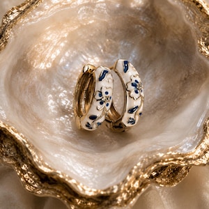 Op de afbeelding: Een paar gouden oorringen met witte emaille bloemmotieven en kleine kristallen accenten. De oorbellen hebben gouden sluitingen en worden gepresenteerd in een goudomrande, schelpvormige schaal.