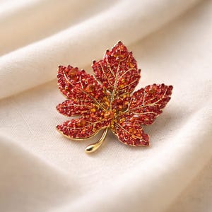 Peut inclure: Broche en forme de feuille d'érable ornée de cristaux rouges et orangés et d'une tige dorée. Accessoire sur le thème de l'automne. La broche est sertie dans un cadre doré.
