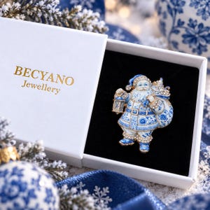 Pode incluir: Um broche de Pai Natal com um padrão floral azul e branco estilo porcelana, detalhes em tom dourado e strass transparentes. O broche é apresentado numa caixa de presente branca com o texto dourado BECYANO Jewellery impresso na tampa.