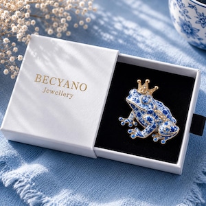 May include: A frog-shaped brooch featuring a blue and white floral pattern with gold-tone accents and a small crown, presented in a white box labelled BECYANO Jewellery. The brooch is set against a black interior lining within the open box.