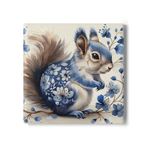 Op de afbeelding: Vierkante keramische tegel met een aquarel illustratie van een eekhoorn. De eekhoorn heeft blauwe en witte bloemmotieven op zijn lichaam en een pluizige bruine staart. Blauwe bloemen en takken omringen de eekhoorn tegen een crèmekleurige achtergrond.