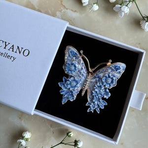 May include: A blue and white butterfly brooch with floral detailing, presented in a white box. The brooch has gold-coloured accents and is set against a black background. The box has the word "CYANO" printed on it.