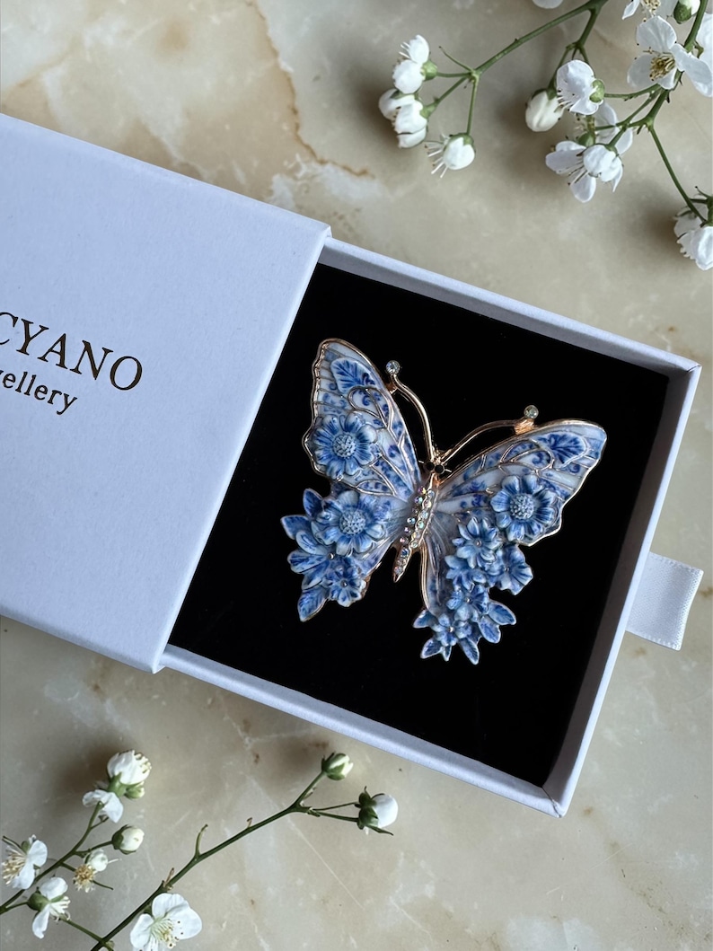May include: A blue and white butterfly brooch with floral detailing, presented in a white box. The brooch has gold-coloured accents and is set against a black background. The box has the word "CYANO" printed on it.