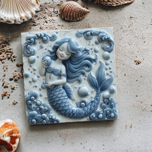 Op de afbeelding: Een blauwe en witte keramische tegel met een 3D-zeemeerminontwerp. De zeemeermin houdt een parel vast en heeft lang, golvend haar. De tegel is omgeven door bellen en golven.