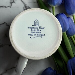 May include: A white ceramic mug with a handle, featuring blue text and a house illustration. The text reads "'t Delftsche Huys' Delft Blue Handwork Made in Holland." The mug is set against a backdrop of blue tulips and a marble surface.