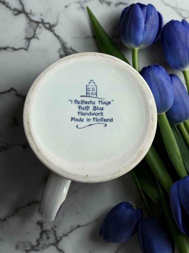 May include: A white ceramic mug with a handle, featuring blue text and a house illustration. The text reads "'t Delftsche Huys' Delft Blue Handwork Made in Holland." The mug is set against a backdrop of blue tulips and a marble surface.