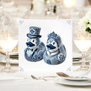 May include: A pair of blue and white ceramic duck figurines dressed as a bride and groom. The groom is wearing a top hat and the bride has a veil. The figurines are on a white background.