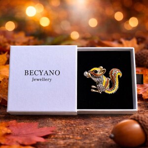 May include: A squirrel brooch with yellow, brown, and silver accents, presented in a white box. The brooch features a pearl and is set against a black background. The box is labeled "BECYANO Jewellery".
