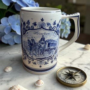 May include: A white ceramic beer stein with blue floral and scenic designs, featuring the word "Heineken." The stein has a large handle and is decorated with a horse-drawn carriage scene. A compass and seashells are also in the image.