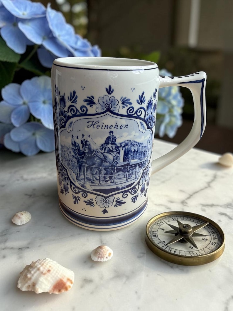 May include: A white ceramic beer stein with blue floral and scenic designs, featuring the word "Heineken." The stein has a large handle and is decorated with a horse-drawn carriage scene. A compass and seashells are also in the image.