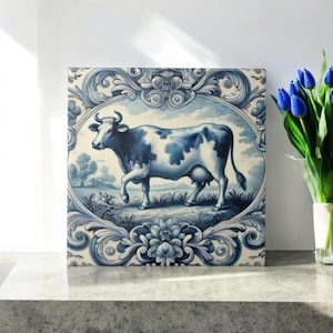 Può includere: Una piastrella di ceramica blu e bianca con un bordo decorativo e una mucca al centro. La mucca è in piedi in un campo con un cielo blu e nuvole bianche sullo sfondo.