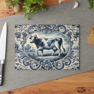 Op de afbeelding: Een blauwe en witte keramische tegel met een decoratieve rand en een koe in het midden. De koe staat op een veld met een boerderij op de achtergrond.