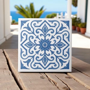 Può includere: Piastrella quadrata bianca con un motivo floreale blu. Il design presenta un fiore centrale circondato da foglie vorticose e forme geometriche. La piastrella è su una superficie di legno, con uno sfondo esterno sfocato.
