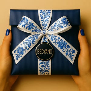 May include: A navy blue gift box tied with a white ribbon adorned with a blue floral pattern. The box is held by hands with blue nail polish. A black and gold circular tag with "BECYANO" is attached to the ribbon.