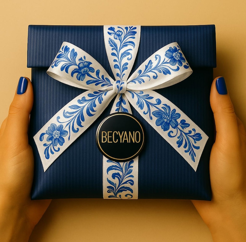 May include: A navy blue gift box tied with a white ribbon adorned with a blue floral pattern. The box is held by hands with blue nail polish. A black and gold circular tag with "BECYANO" is attached to the ribbon.