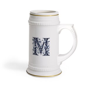 Op de afbeelding: Witte keramische bierpul met een gouden rand en handvat. De pul heeft een decoratieve blauw-witte letter "M" in een sierlijk bloemmotief. De pul heeft een getextureerd oppervlak en een klassieke vorm.