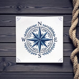 May include: Square white tile with a blue compass rose design. The compass rose is surrounded by a wreath and the cardinal directions are labeled. Decorative floral accents are in each corner. A rope is in the background.