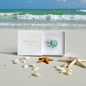 Könnte beinhalten: Eine geöffnete weiße Schachtel mit dem Text "BECYANO Jewellery" zeigt eine türkis-weiße Muschelbrosche. Die Brosche liegt auf einer weißen Oberfläche, umgeben von Muscheln und Seesternen. Der Hintergrund zeigt das Meer und den Himmel.